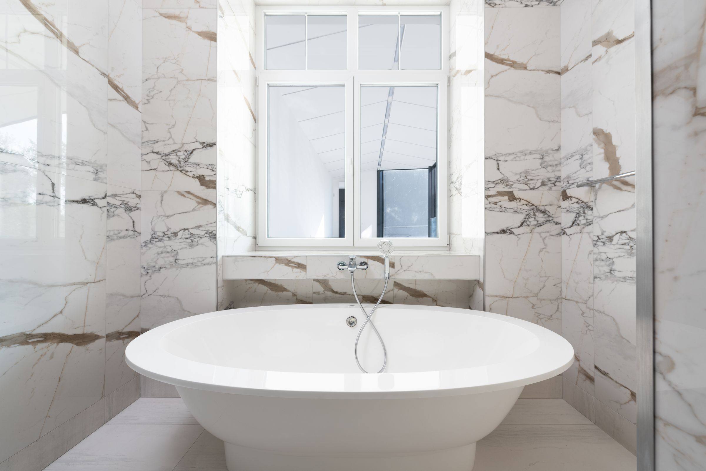 Marble-look tiled bathroom with a freestanding tub beneath a window