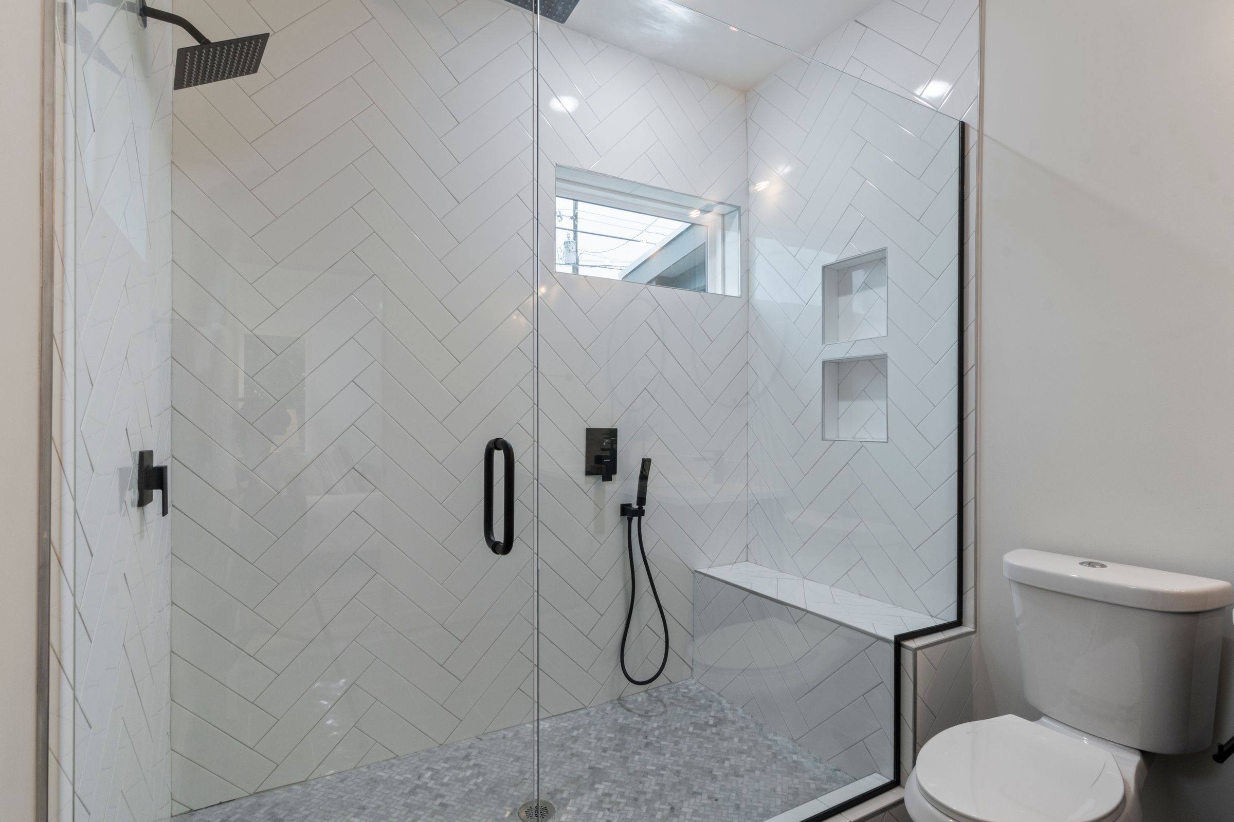 White herringbone-tile walk-in shower with a glass enclosure, bench, niches and black fixtures